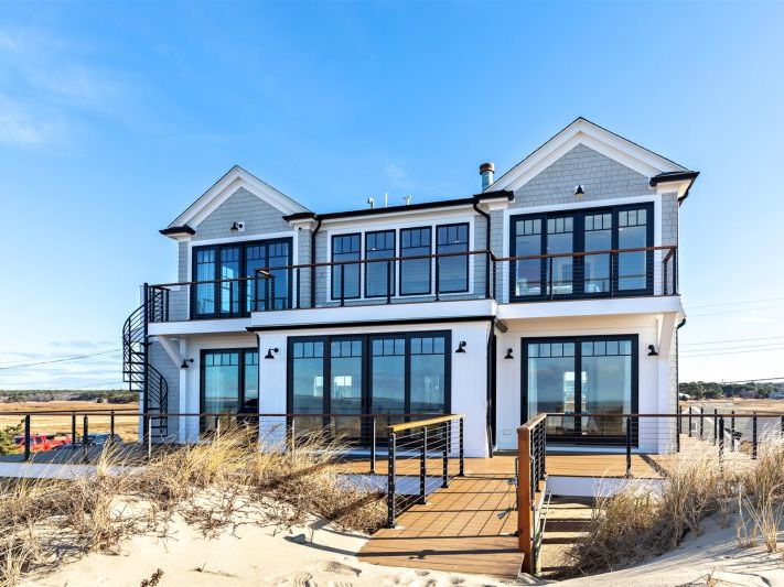 Modern coastal home with gray shingle siding, expansive glass doors and balconies, a spiral staircase to the upper deck, and a private boardwalk leading through the dunes under a clear blue sky.