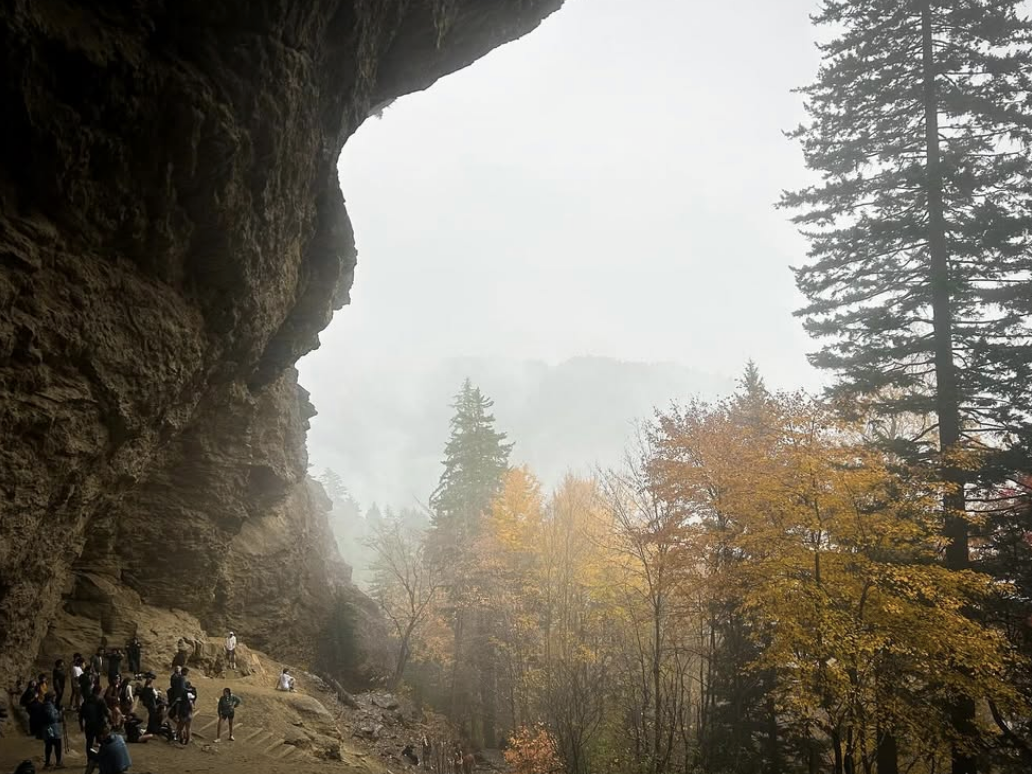 Hikers gather beneath the overhanging rock formation at Alum Cave Bluffs in the Great Smoky Mountains, surrounded by misty fall foliage and mountain views in the distance.