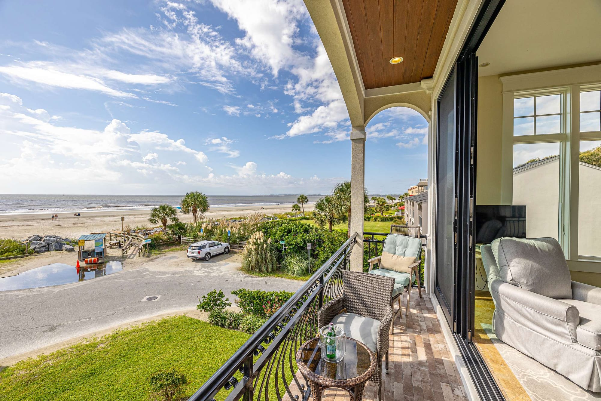 A private balcony at this oceanfront vacation rental offers sweeping views of the beach, dunes, and Atlantic Ocean, furnished with wicker seating beneath a covered wood-paneled archway. The adjoining living room with a plush sofa flows seamlessly onto the outdoor space, blending indoor comfort with direct coastal access.