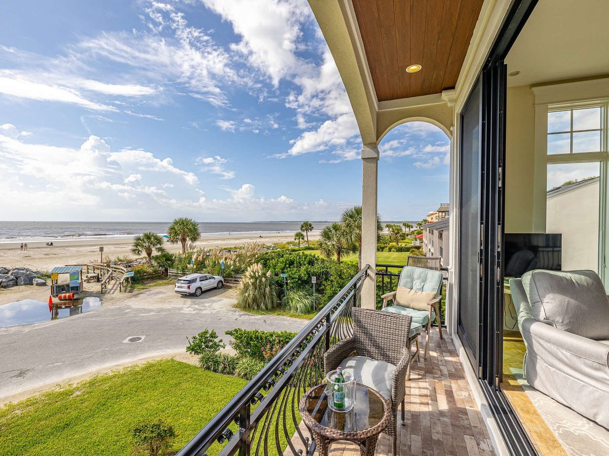 A private balcony at this oceanfront vacation rental offers sweeping views of the beach, dunes, and Atlantic Ocean, furnished with wicker seating beneath a covered wood-paneled archway. The adjoining living room with a plush sofa flows seamlessly onto the outdoor space, blending indoor comfort with direct coastal access.