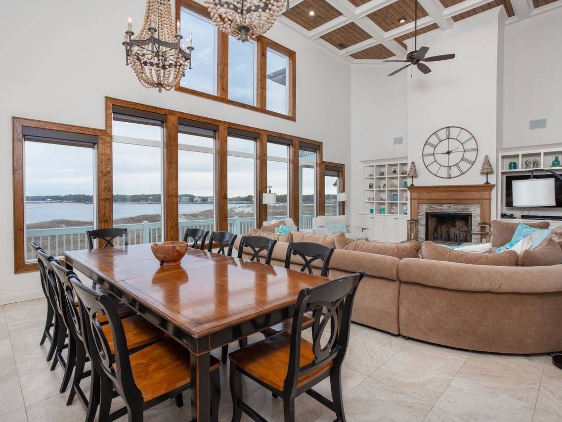 A spacious coastal living and dining area at Holden Beach with tall windows overlooking the water. The room features a large wooden dining table, cozy sectional sofa, chandelier lighting, and a stone fireplace for gatherings.