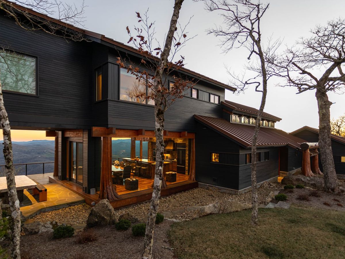 The Flying Eagle at Eagles Nest is a modern mountain retreat surrounded by trees with wide windows that look out across the Blue Ridge Mountains. Its dark exterior and warm interior lighting create a cozy setting for a peaceful mountain escape.