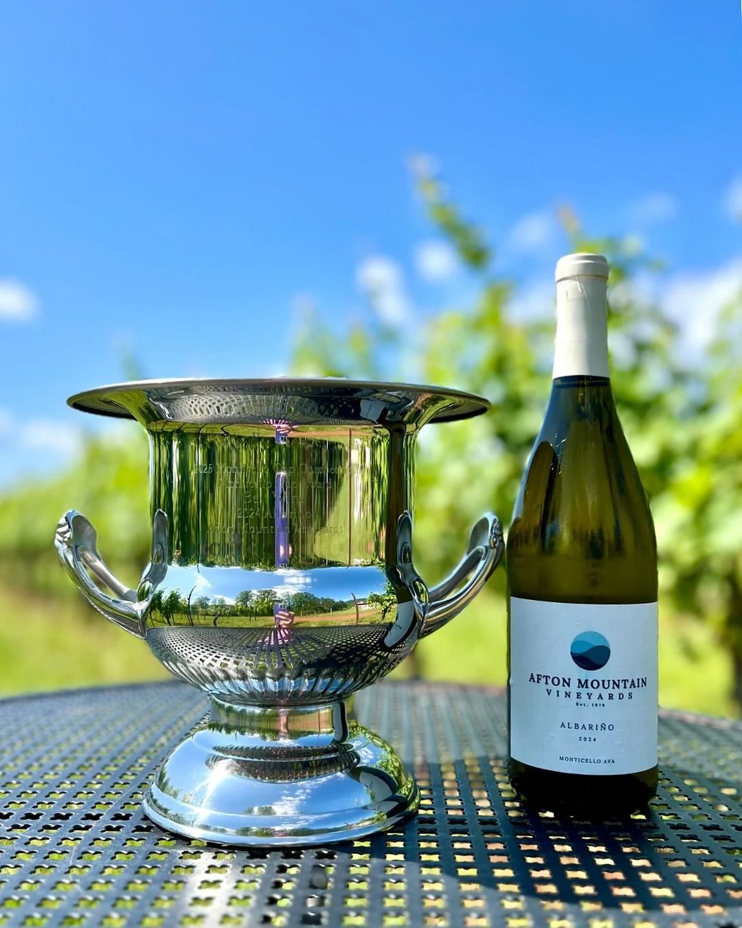 A chilled bottle of Albariño wine from Afton Mountain Vineyards stands next to a silver trophy under a bright blue sky. The polished setup and vineyard reflection celebrate excellence in Monticello’s award-winning wines.