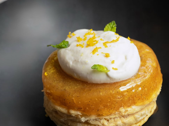 A layered pastry topped with caramelized fruit, a dollop of whipped cream, fresh mint, and orange zest served on a black plate.