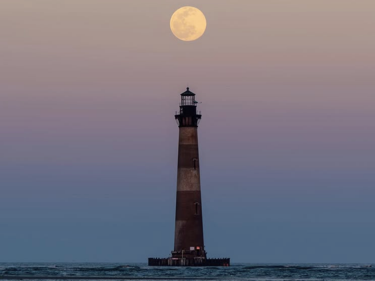 A solitary lighthouse stands in the ocean beneath a full moon at dusk, silhouetted against a gradient sky of soft purples and blues.