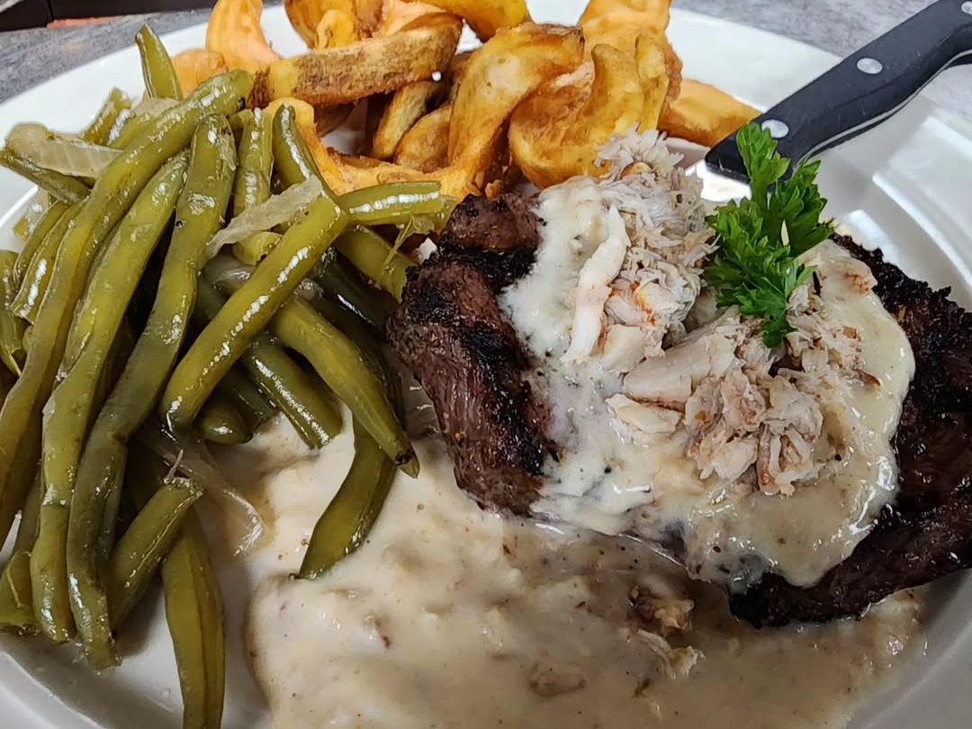 A grilled steak is topped with creamy crab sauce and served with crisp green beans and golden fries. White Horse Tavern offers a hearty and indulgent dish that captures classic American flavors.