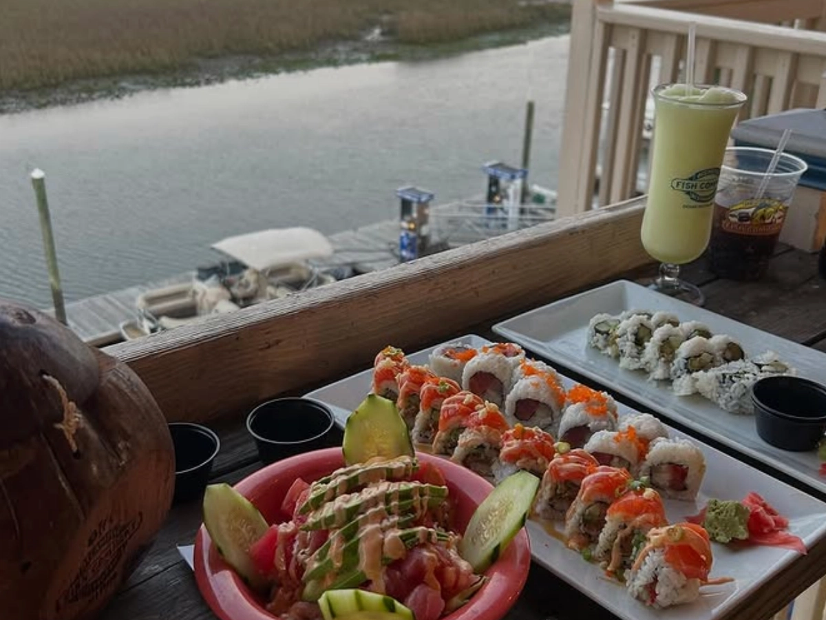 A colorful sushi spread and poke bowl served on a wooden deck overlooking a serene marsh and docked boats, with tropical drinks and dipping sauces on the side.