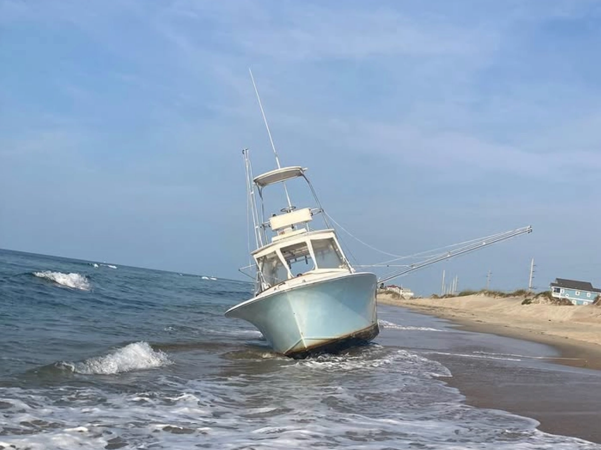 A fishing boat leans on its side, grounded in the surf along a sandy beach under a clear blue sky.