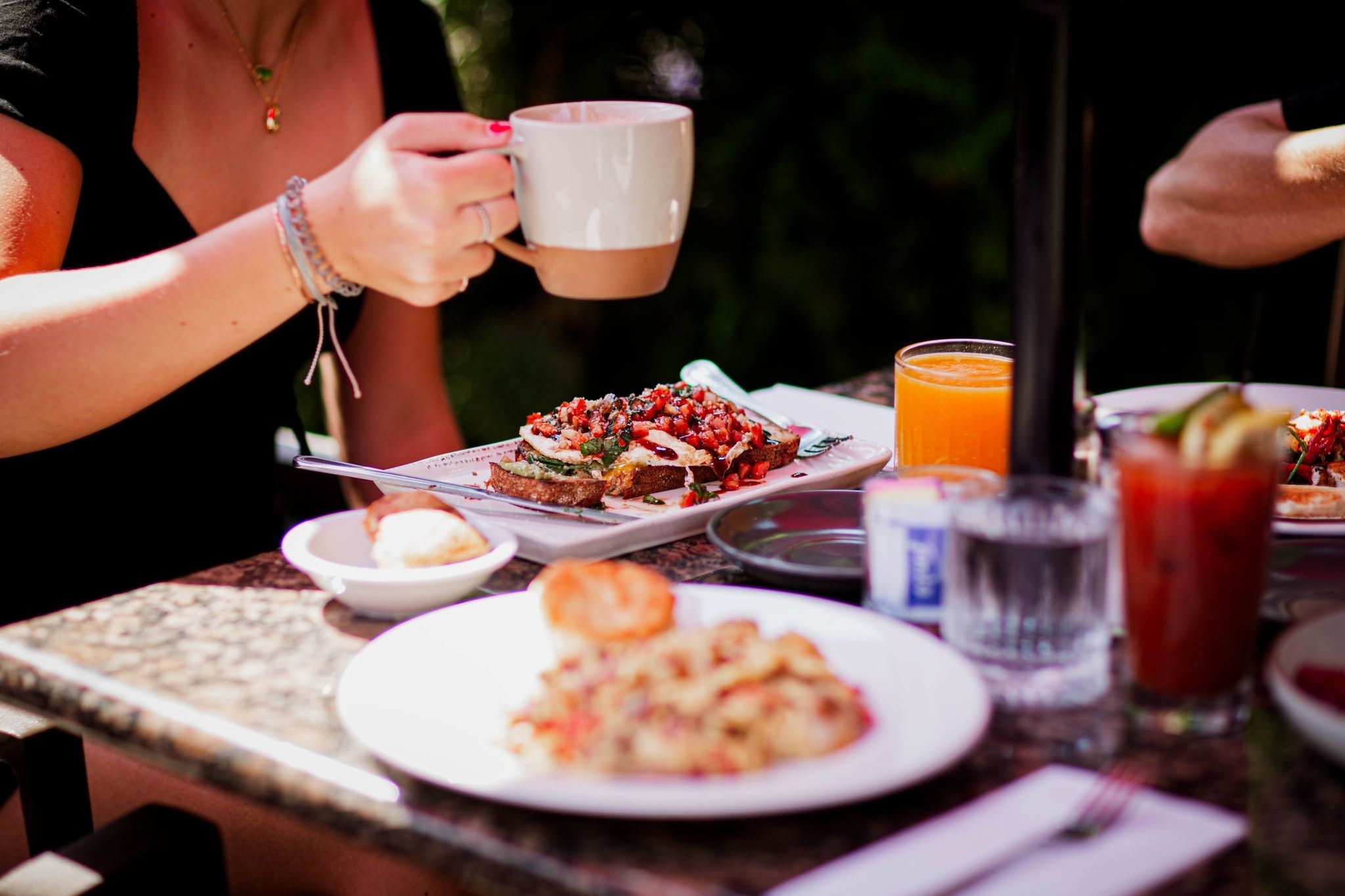 A cozy outdoor brunch spread features avocado toast topped with bruschetta, a plate of home fries with a biscuit, fresh orange juice, and Bloody Marys on a marble-topped table. A woman sips coffee from a ceramic mug amid the relaxed, sunlit garden setting.