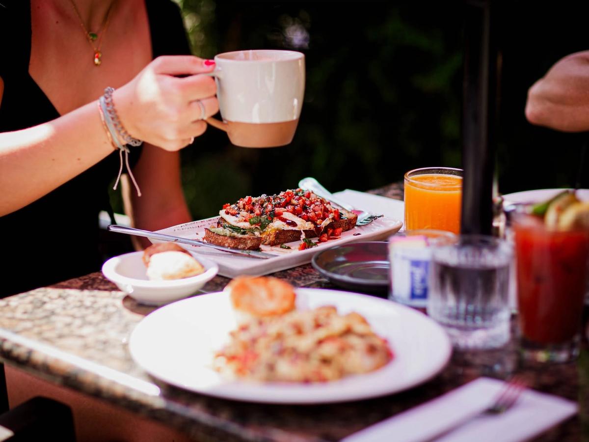 A cozy outdoor brunch spread features avocado toast topped with bruschetta, a plate of home fries with a biscuit, fresh orange juice, and Bloody Marys on a marble-topped table. A woman sips coffee from a ceramic mug amid the relaxed, sunlit garden setting.