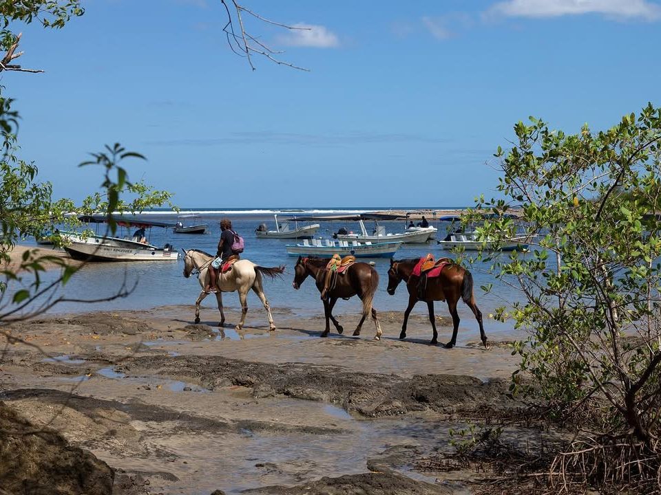 Person riding a horse along the shore with two more horses, near anchored boats in a calm bay. A unique mix of beach and horseback adventure in Costa Rica.