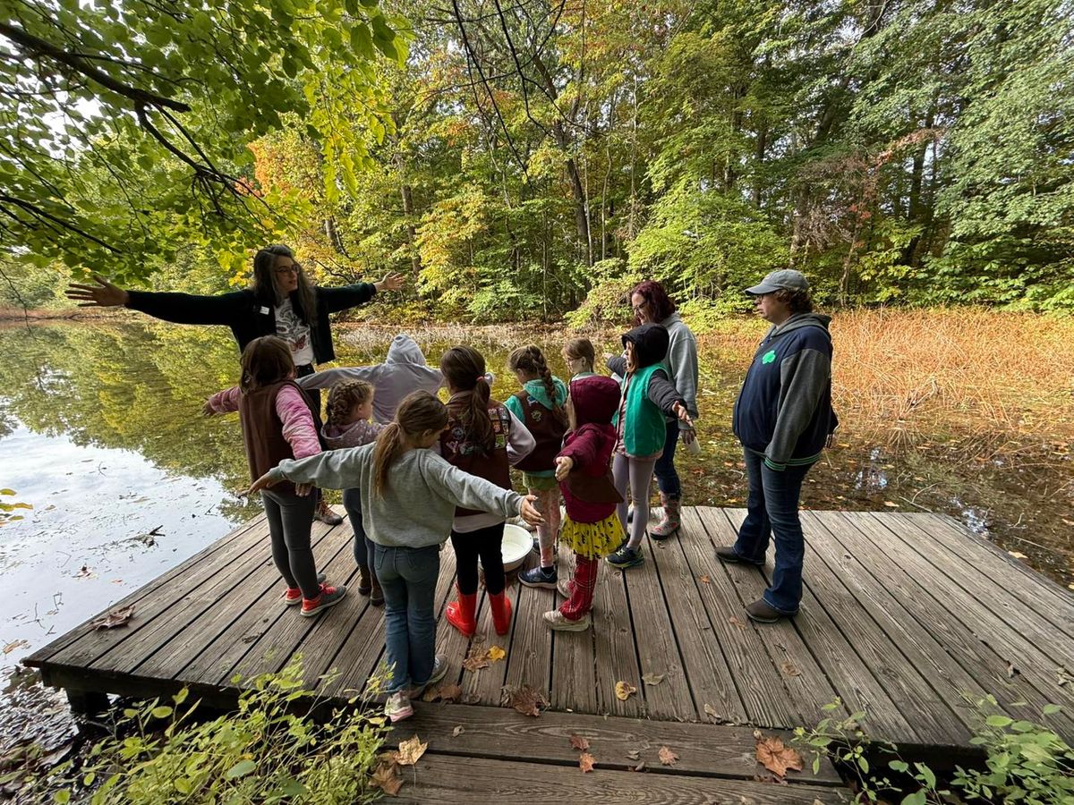 Children and adults gather on a wooden dock surrounded by lush greenery and calm pond waters, learning about nature in a hands-on way. The peaceful forest backdrop makes it the perfect place for outdoor exploration and discovery.