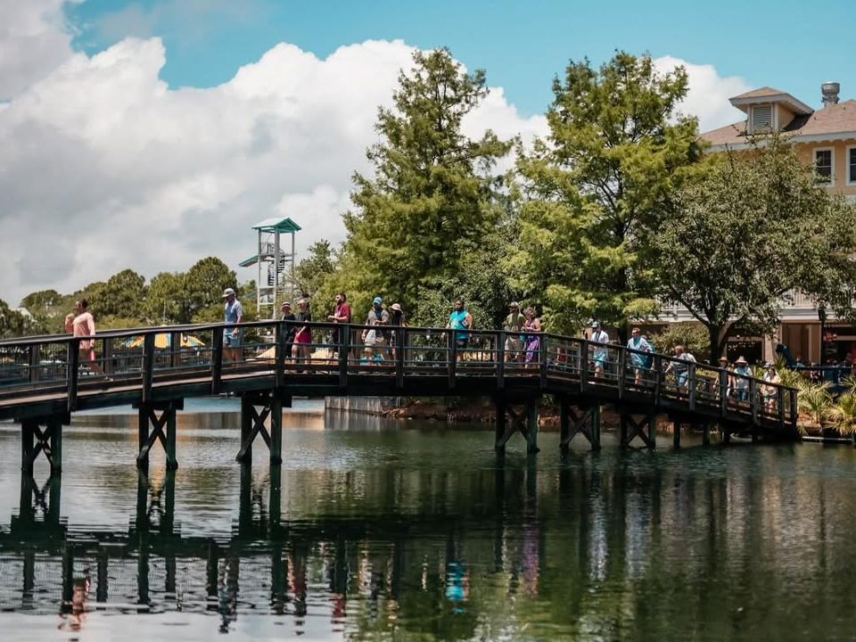 Visitors crossing a wooden bridge at Baytowne Wharf surrounded by water, trees, and shops, creating a lively village vibe in Sandestin.