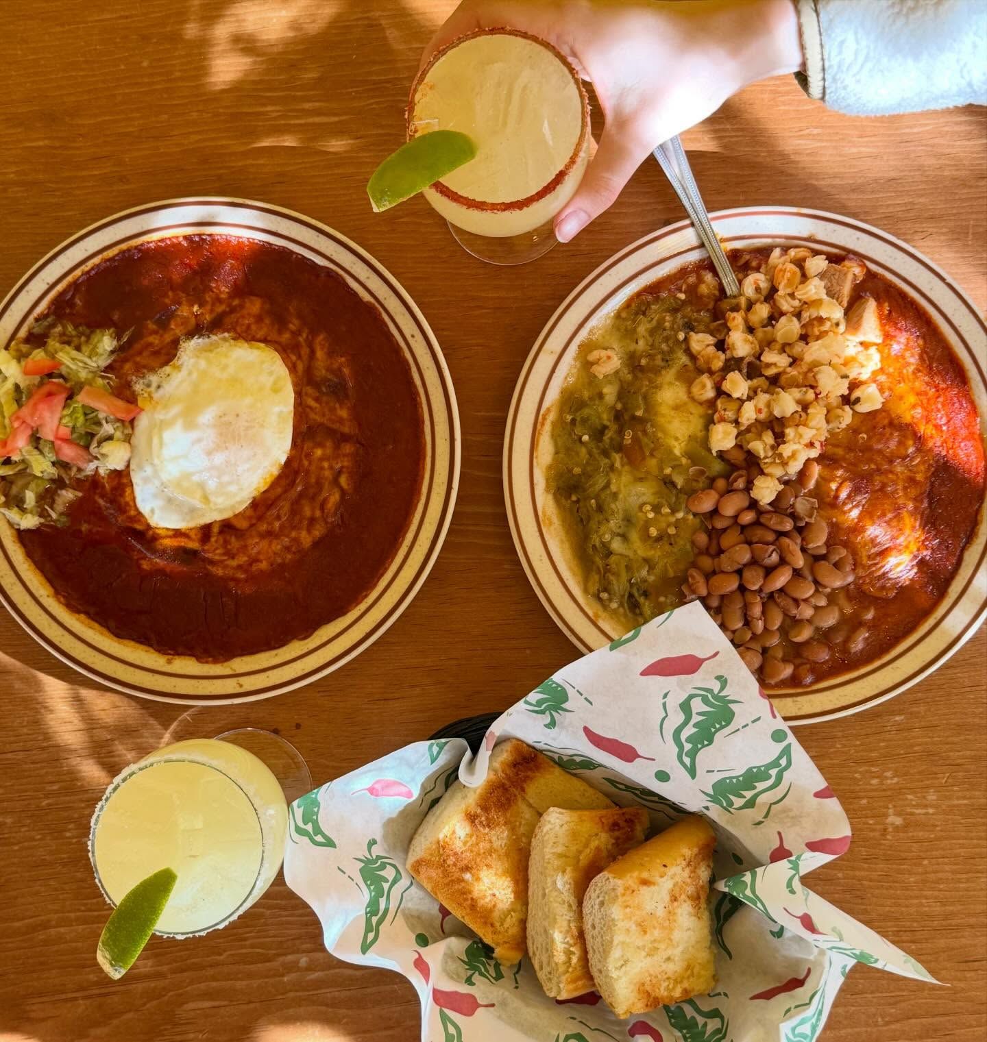 An overhead view of two red chile plates topped with a fried egg and posole, served with pinto beans and a basket of chile-dusted bread rolls on a warm wooden table. Two frozen margaritas with lime wedges round out the sunlit New Mexican meal.