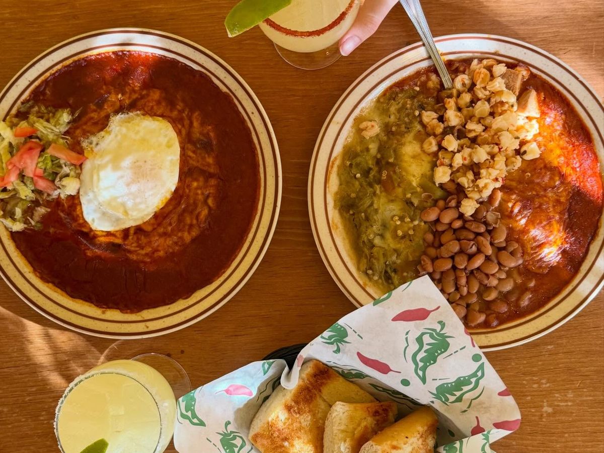 An overhead view of two red chile plates topped with a fried egg and posole, served with pinto beans and a basket of chile-dusted bread rolls on a warm wooden table. Two frozen margaritas with lime wedges round out the sunlit New Mexican meal.