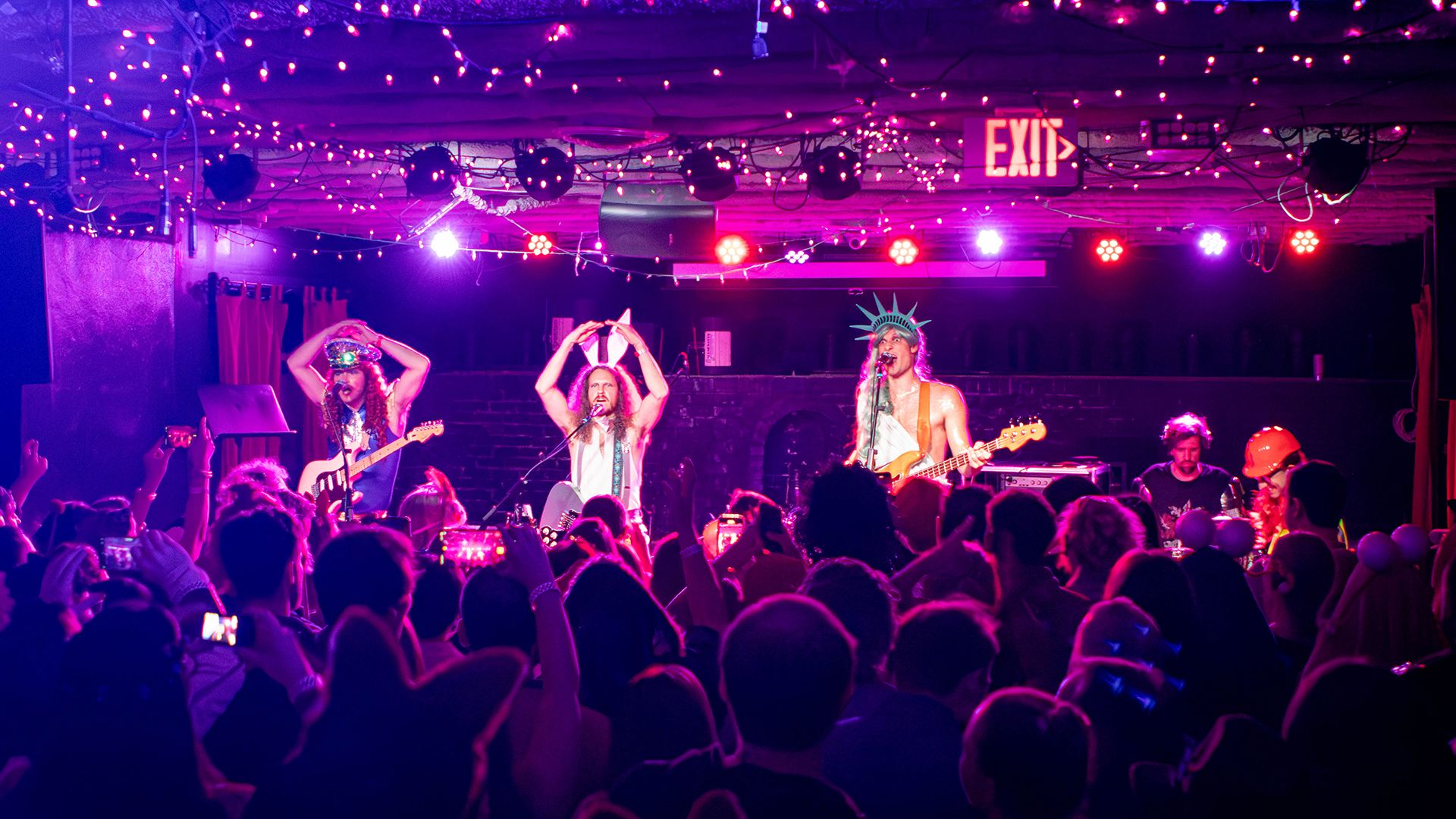 A packed crowd fills a dimly lit music venue bathed in pink and purple stage lighting as a costumed band performs on stage. String lights line the ceiling above the energetic crowd, who are filming the show on their phones.
