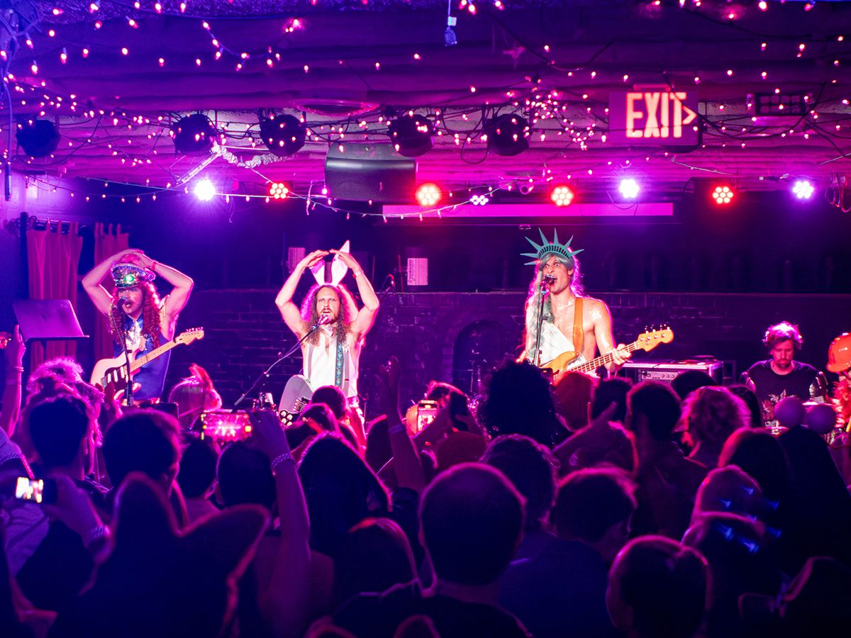 A packed crowd fills a dimly lit music venue bathed in pink and purple stage lighting as a costumed band performs on stage. String lights line the ceiling above the energetic crowd, who are filming the show on their phones.