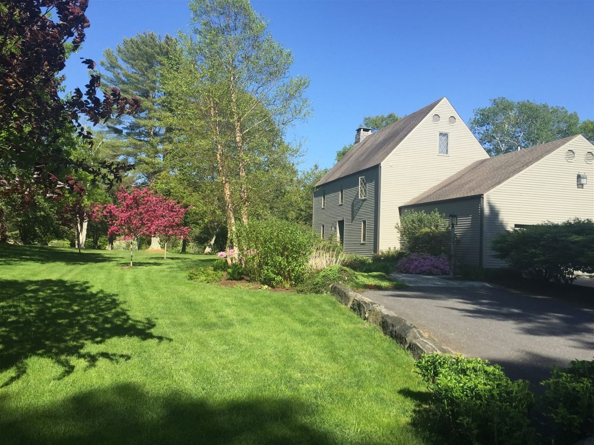 A light-gray house with a steep roof sits on a well-kept lawn, surrounded by vibrant trees and flowering bushes under a bright blue sky.