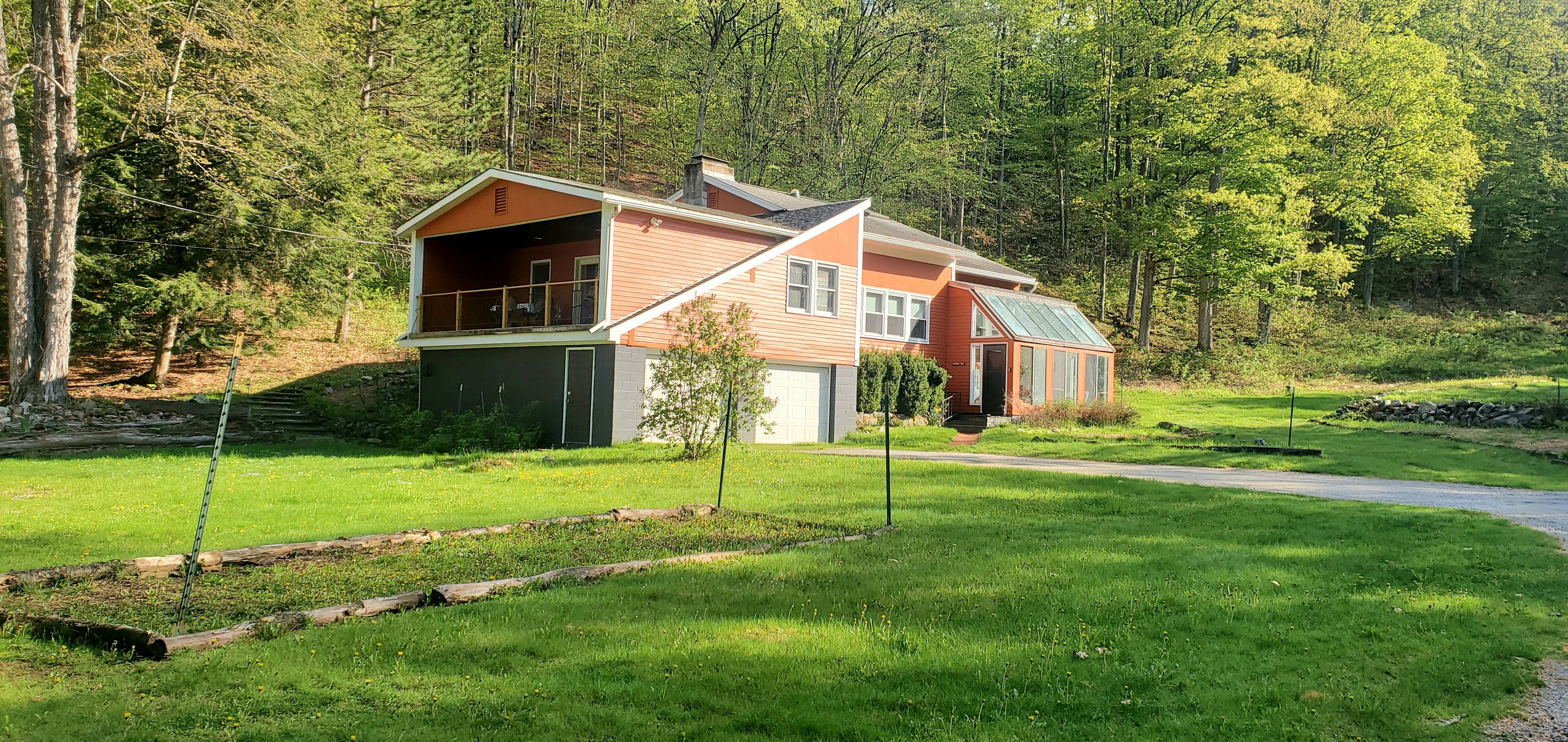 A unique orange and gray house with a covered upper deck and a sunroom-style glass entryway, set against a lush green lawn and surrounded by tall trees on a sunny day.