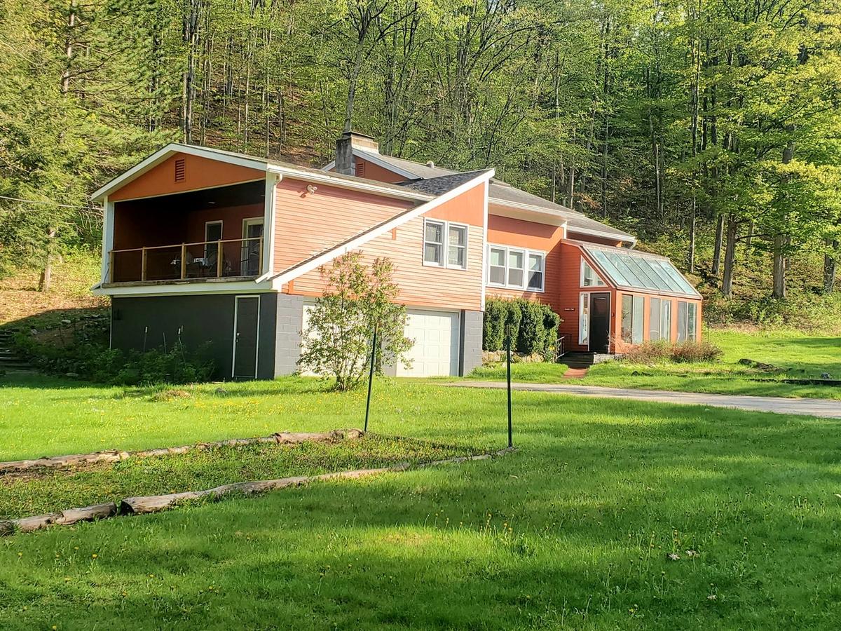 A unique orange and gray house with a covered upper deck and a sunroom-style glass entryway, set against a lush green lawn and surrounded by tall trees on a sunny day.