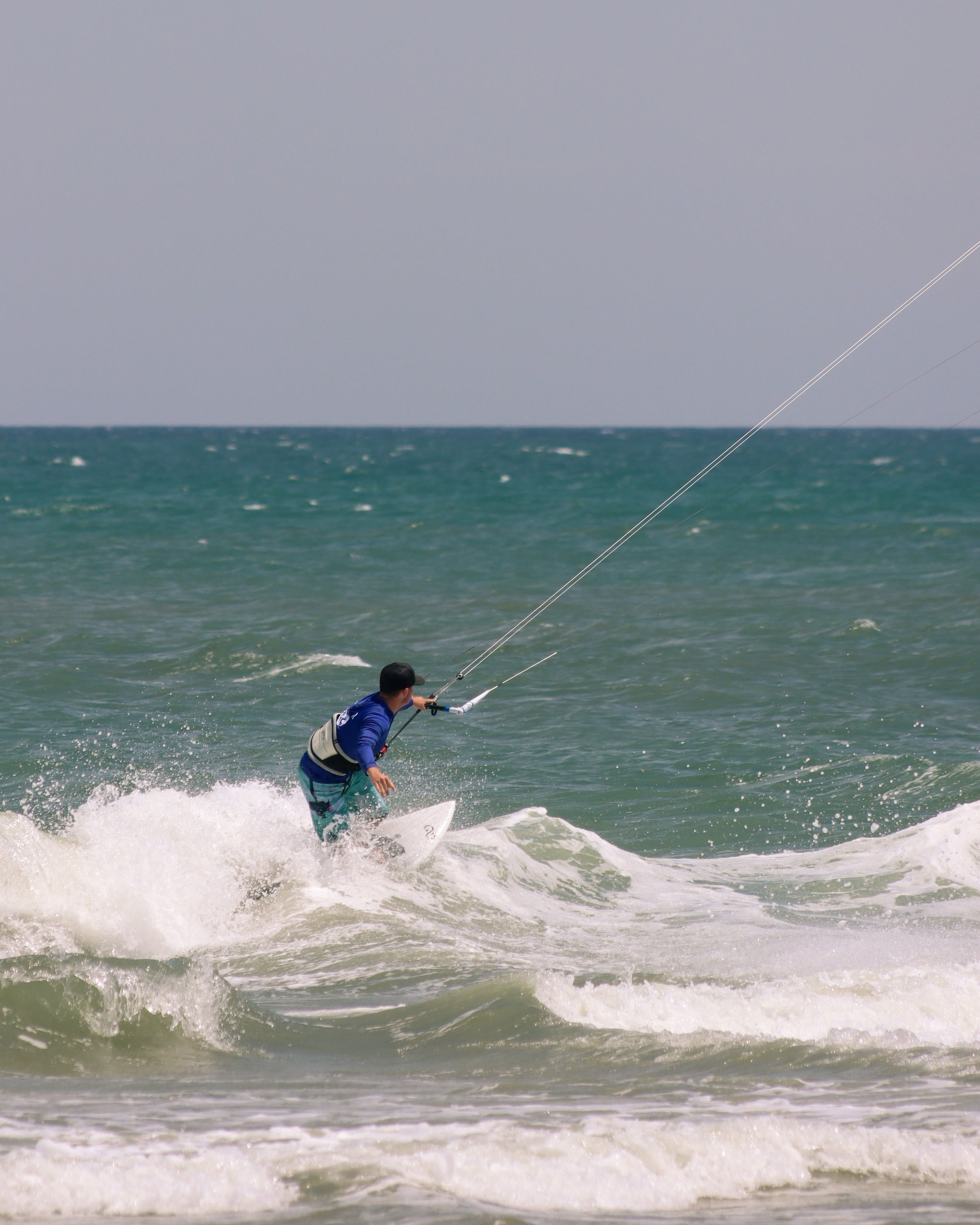 A kiteboarder in a blue rash guard and life vest carves across a breaking wave, gripping the control bar as lines stretch up toward the sky. The teal ocean churns with whitewater under a pale, hazy horizon. 