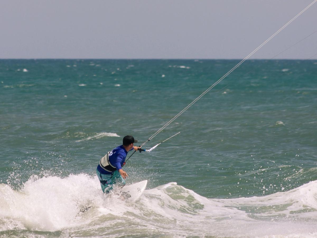 A kiteboarder in a blue rash guard and life vest carves across a breaking wave, gripping the control bar as lines stretch up toward the sky. The teal ocean churns with whitewater under a pale, hazy horizon.