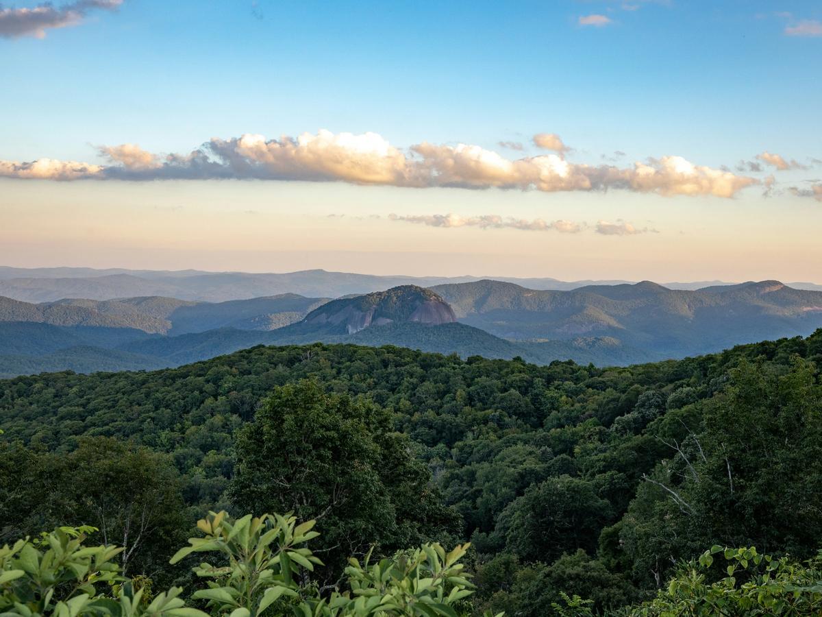 View of Looking Glass Rock surrounded by lush green forests and rolling mountains under a partly cloudy sky in the Blue Ridge Mountains of North Carolina.