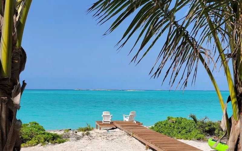 A sandy path with a wooden walkway leads to two white chairs overlooking turquoise waters, framed by palm trees and tropical greenery.