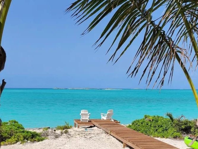 A sandy path with a wooden walkway leads to two white chairs overlooking turquoise waters, framed by palm trees and tropical greenery.