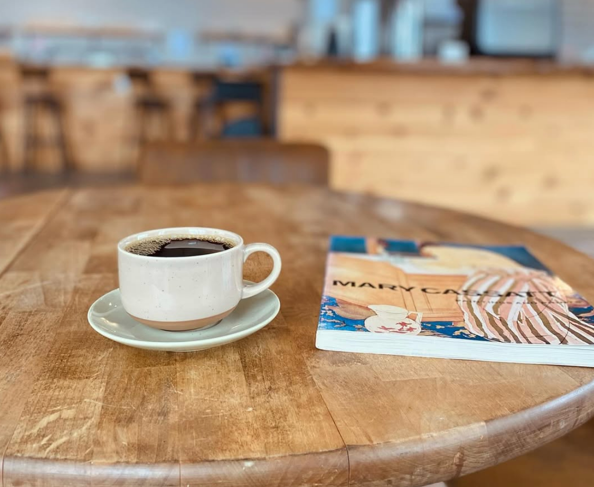 A ceramic cup of black coffee sits on a wooden café table beside a paperback book by Mary Oliver, with a softly blurred, warm-toned coffee shop interior in the background.