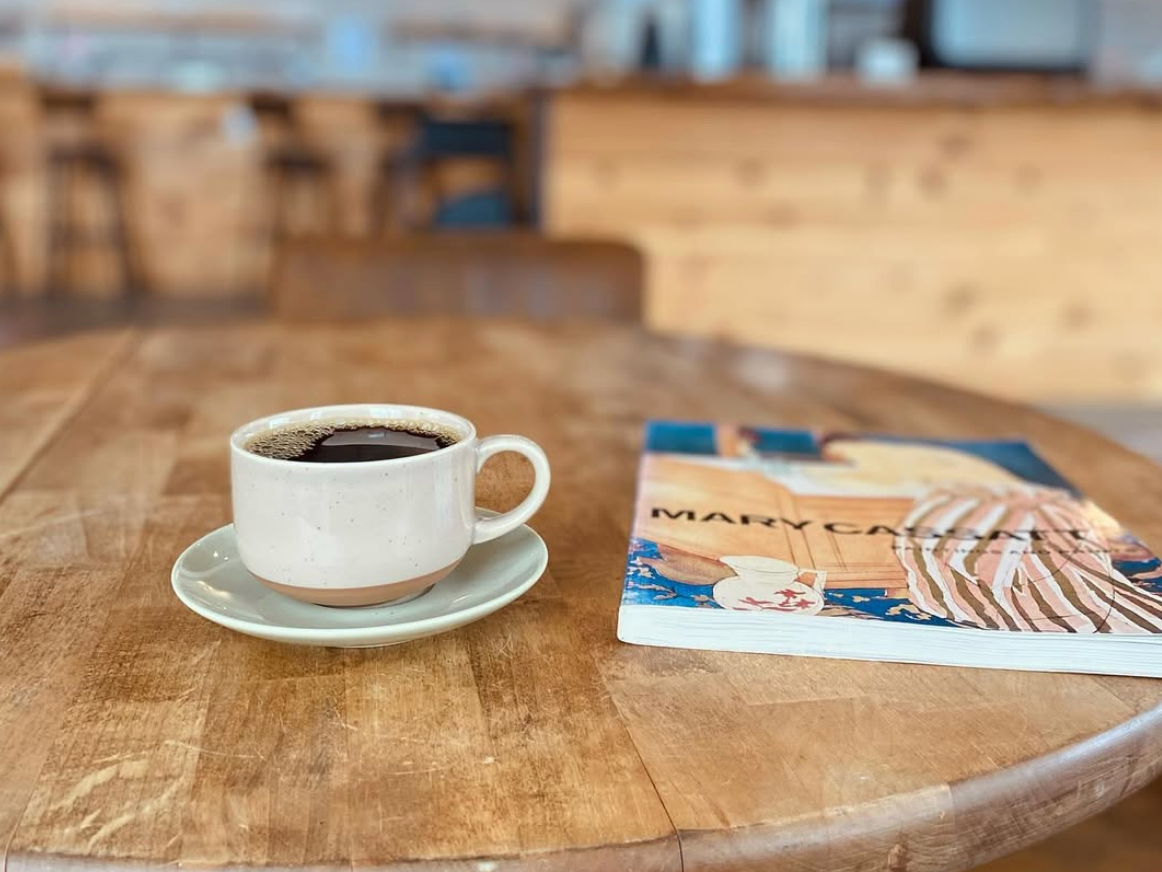 A ceramic cup of black coffee sits on a wooden café table beside a paperback book by Mary Oliver, with a softly blurred, warm-toned coffee shop interior in the background.