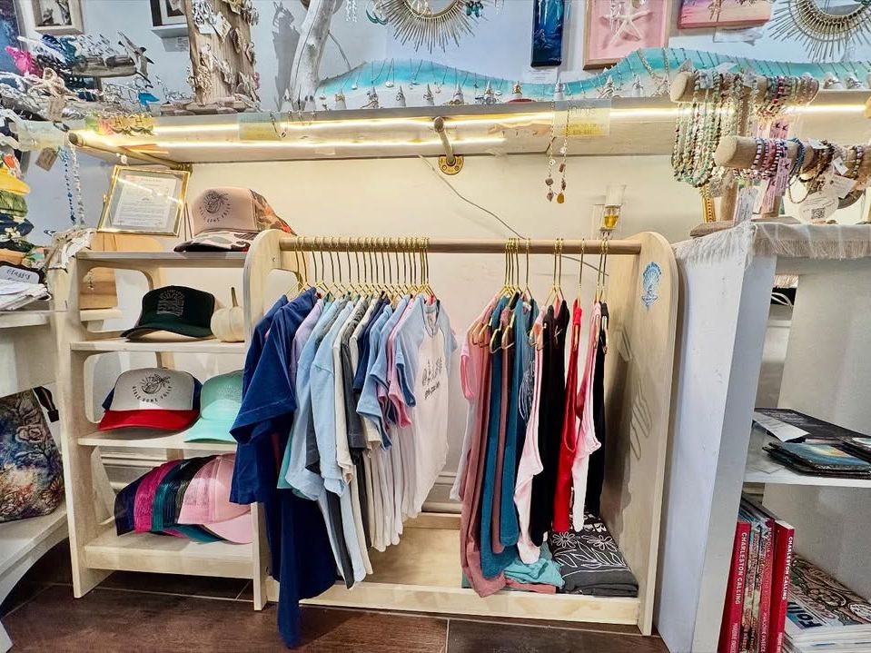 A cozy beach boutique filled with colorful t-shirts, hats, and local jewelry on display. Visitors browse relaxed, coastal-style clothing and souvenirs. Shops like this are perfect for picking up Folly Beach gifts and beachwear.