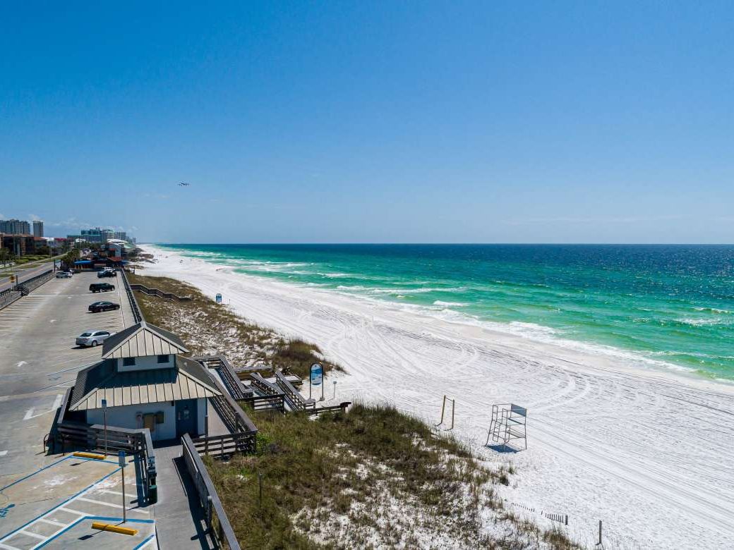Aerial view of Miramar Beach Regional Access showing the wide stretch of white sandy beach, clear blue water, and parking right along the shoreline for easy access.