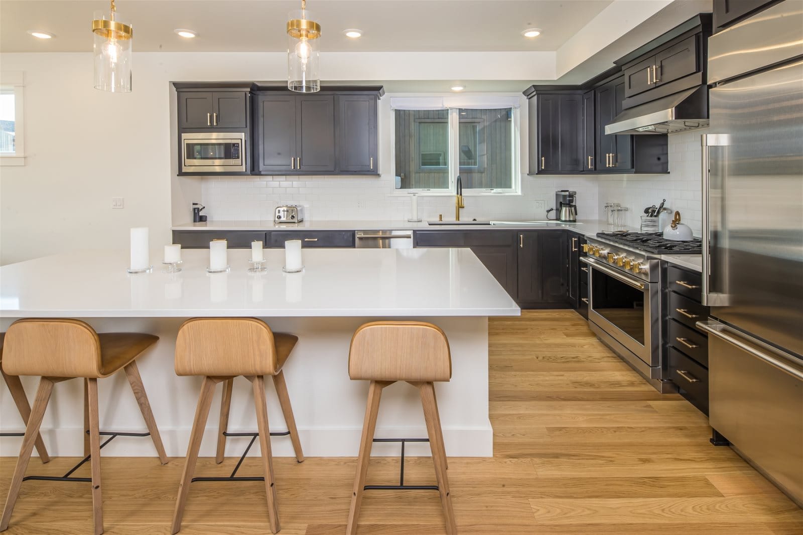 A modern kitchen with dark cabinetry, white countertops, and stainless steel appliances features a large island with wooden barstools and pendant lighting.