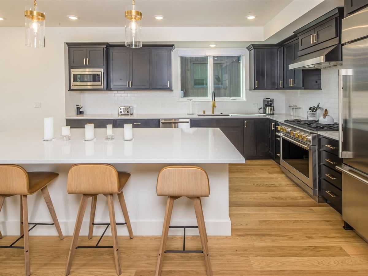 A modern kitchen with dark cabinetry, white countertops, and stainless steel appliances features a large island with wooden barstools and pendant lighting.
