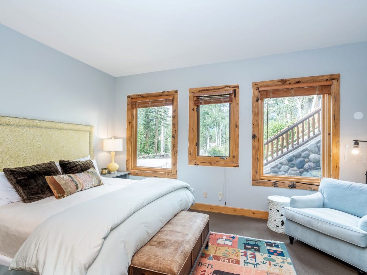 Bright bedroom with a yellow headboard bed, wooden-framed windows, blue armchair, and patterned rug.