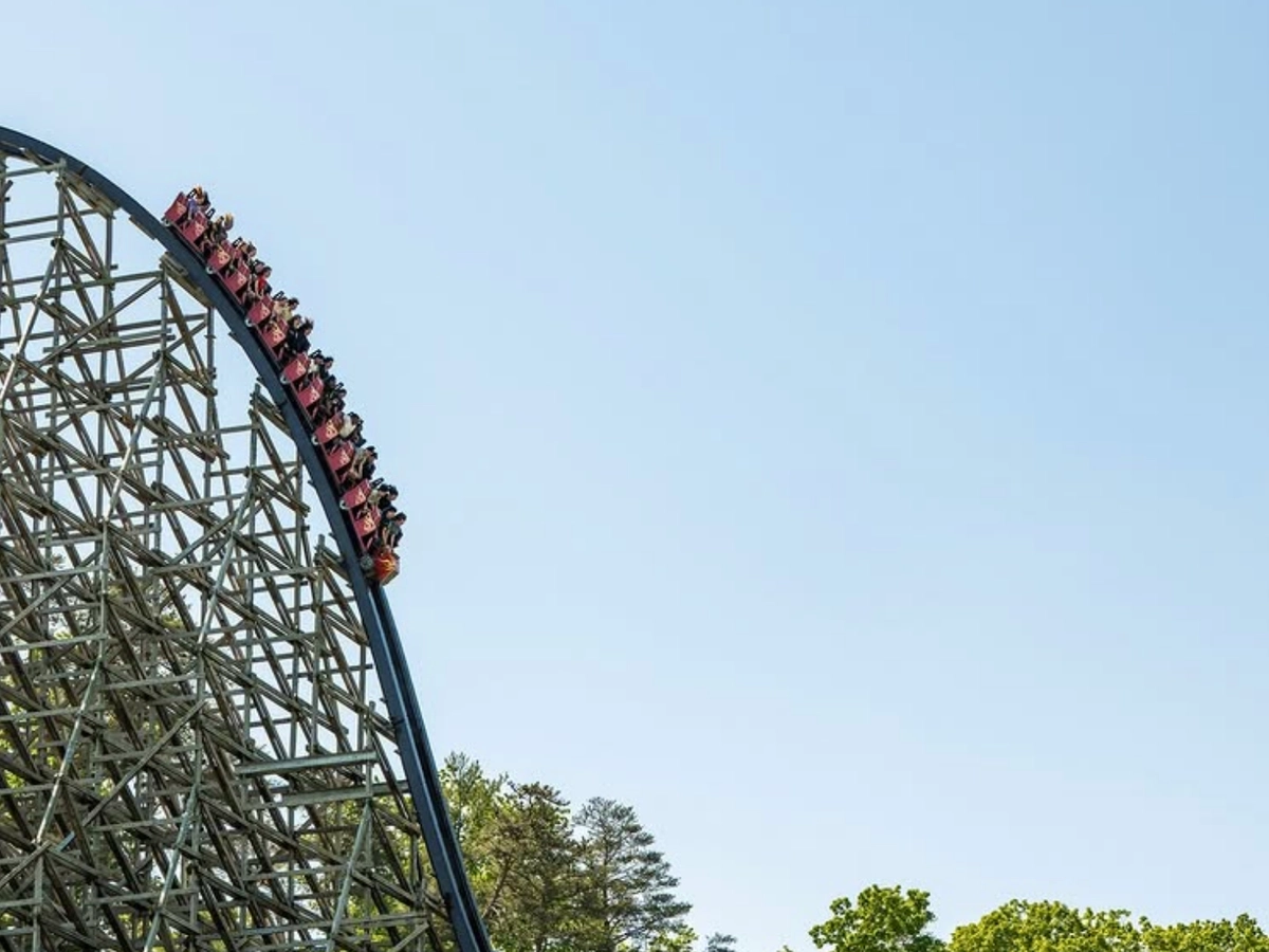 A roller coaster full of riders plunges down a steep wooden track against a clear blue sky.