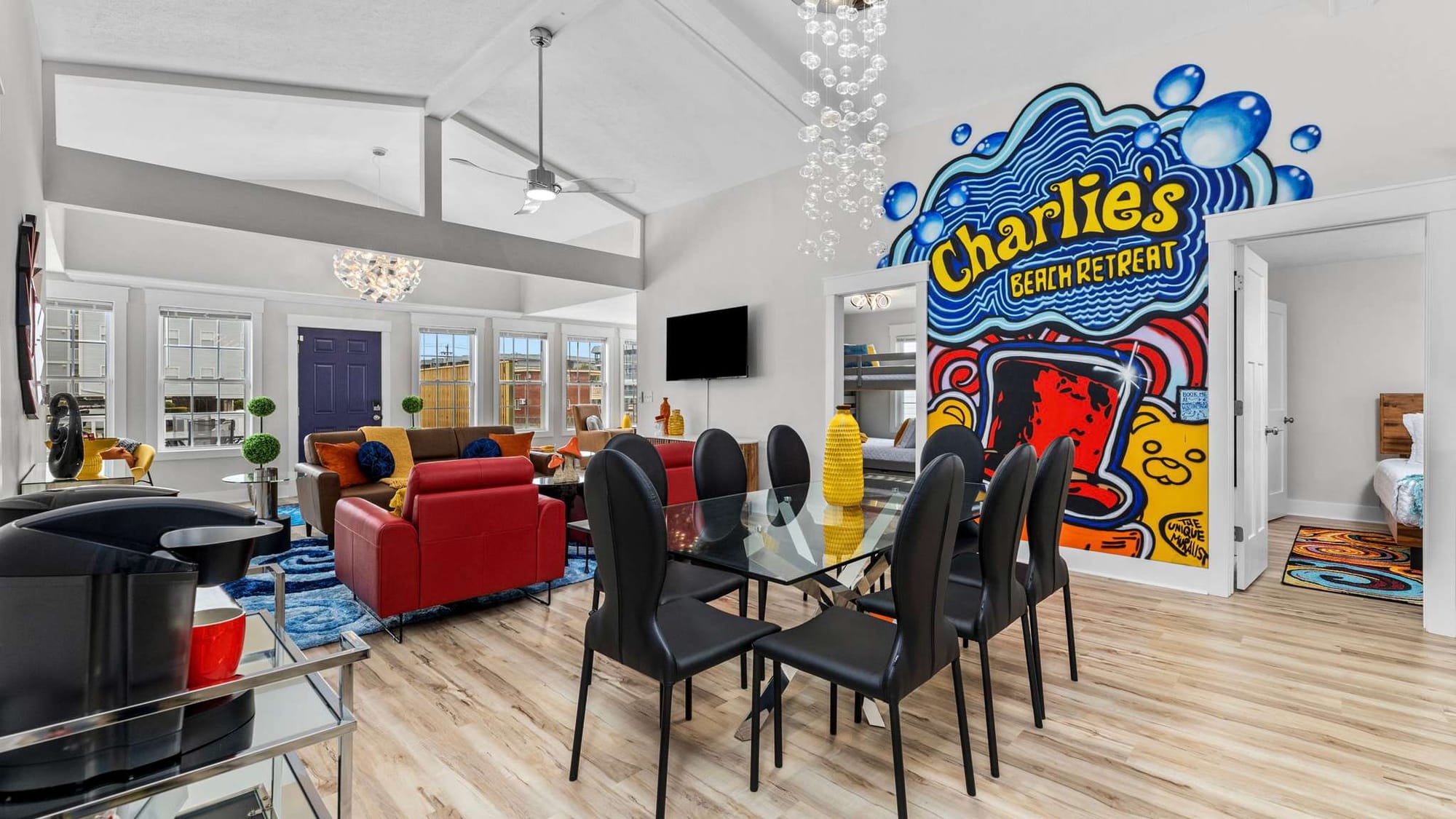 Bright open-concept living and dining space with colorful modern furniture, glass dining table, vaulted ceilings, and a bold “Charlie’s Beach Retreat” mural wall.