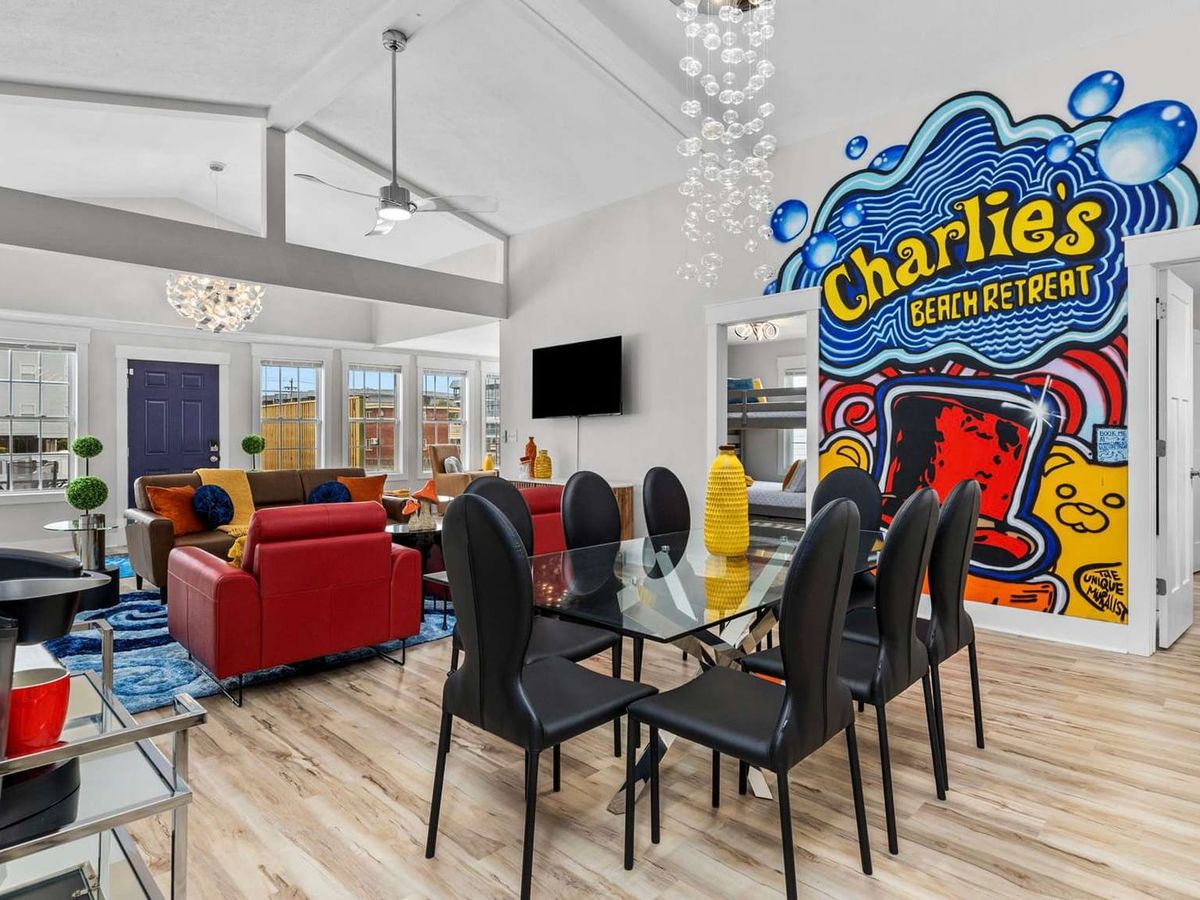 Bright open-concept living and dining space with colorful modern furniture, glass dining table, vaulted ceilings, and a bold “Charlie’s Beach Retreat” mural wall.