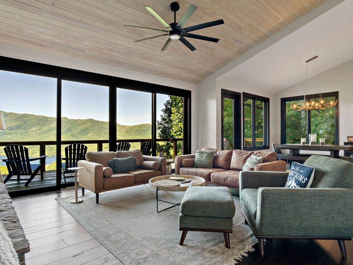 Cozy mountain cabin living room with a tall stone fireplace, leather chair, and big windows showing the trees outside. This peaceful lodge is great for relaxing by the fire after a day of hiking or lake fun.