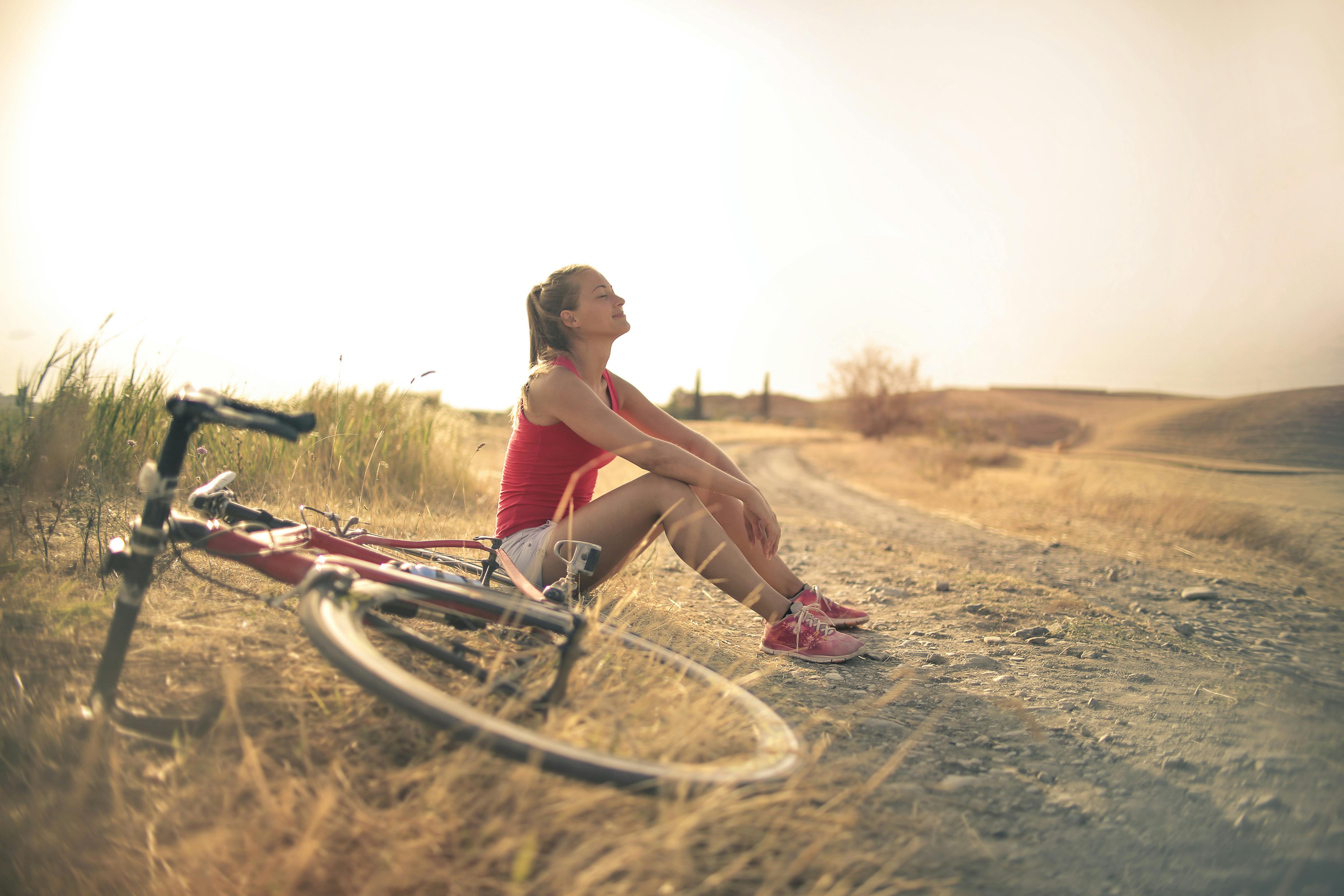 A woman rests beside her bicycle on a quiet dirt road, eyes closed and smiling in the warm light. The open fields and empty path create a calm, mindful moment that feels like a fresh start.