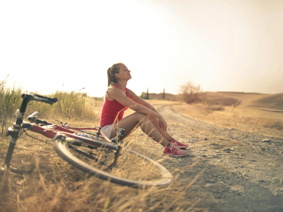 A woman rests beside her bicycle on a quiet dirt road, eyes closed and smiling in the warm light. The open fields and empty path create a calm, mindful moment that feels like a fresh start.