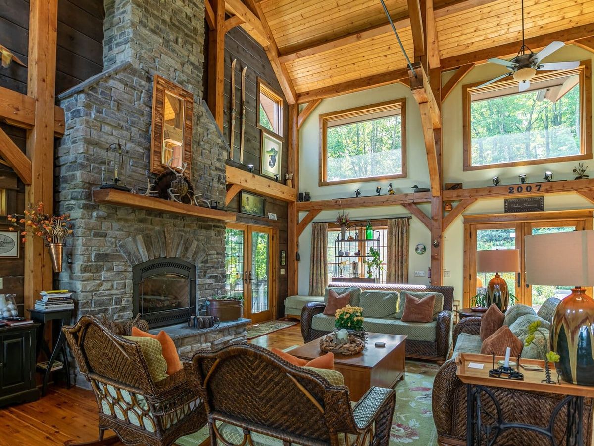 Inside a timber-frame cabin, a stone fireplace rises to the vaulted ceiling while sunlight pours through large windows. Comfortable seating and rustic details create a welcoming space for family gatherings.