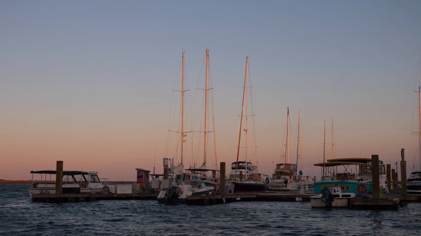 Sailboats and small craft rest quietly at a wooden marina dock as the sky fades into soft peach and blue tones at dusk. Tall masts stand silhouetted against the fading light over the calm harbor water. 