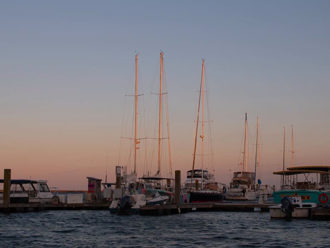 Sailboats and small craft rest quietly at a wooden marina dock as the sky fades into soft peach and blue tones at dusk. Tall masts stand silhouetted against the fading light over the calm harbor water.