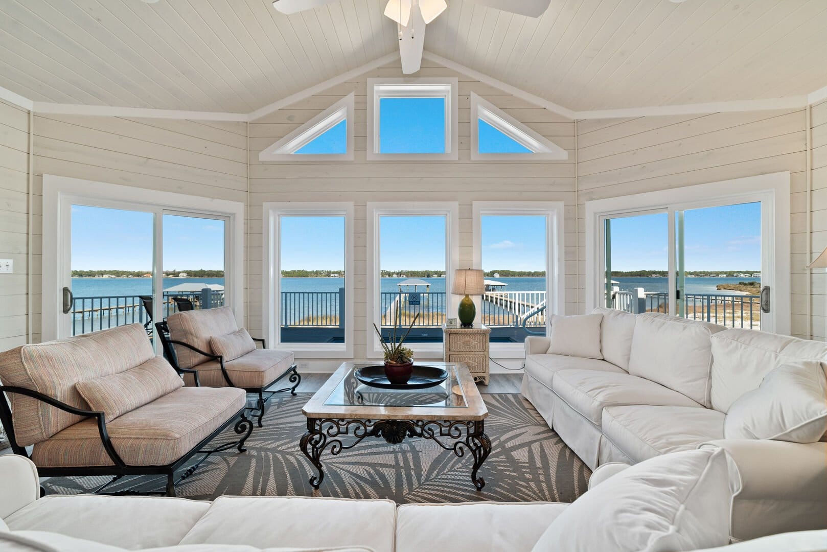 Bright coastal living room with vaulted ceilings, white shiplap walls, and large windows showcasing water views. Furnished with cushioned seating, a glass-top coffee table, and access to the balcony.