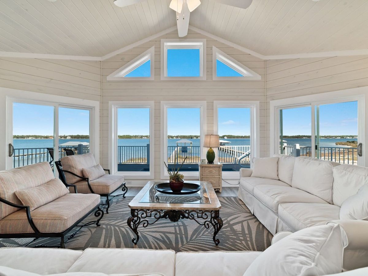 Bright coastal living room with vaulted ceilings, white shiplap walls, and large windows showcasing water views. Furnished with cushioned seating, a glass-top coffee table, and access to the balcony.