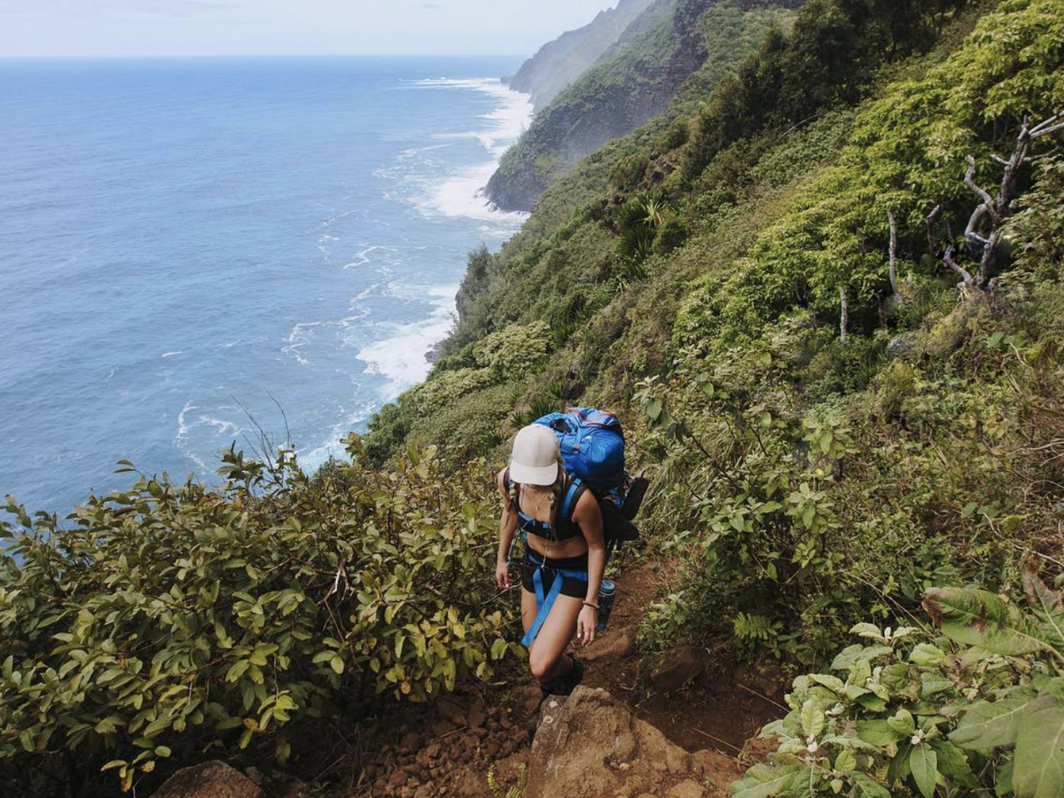 A hiker with a large blue backpack climbs a steep trail along Kauai’s lush Nā Pali Coast, with dramatic green cliffs dropping to the ocean below.