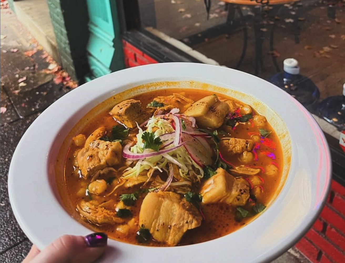 A bowl of hearty chicken soup in a rich red broth with hominy, topped with shredded cabbage, red onion, and cilantro.