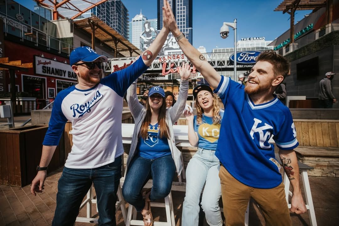 Fans wearing Kansas City Royals gear celebrate with high-fives and laughter in an energetic downtown setting. The bright day and lively atmosphere capture the excitement of sports and community spirit.